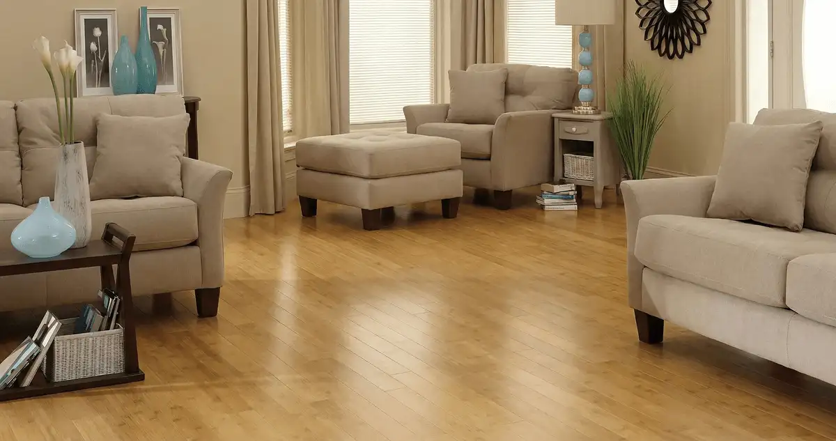 A living room with professionally installed bamboo flooring, beige sofas, a matching ottoman, a side table with a blue lamp, vases, framed pictures, and large windows with blinds and curtains in Toledo, OH